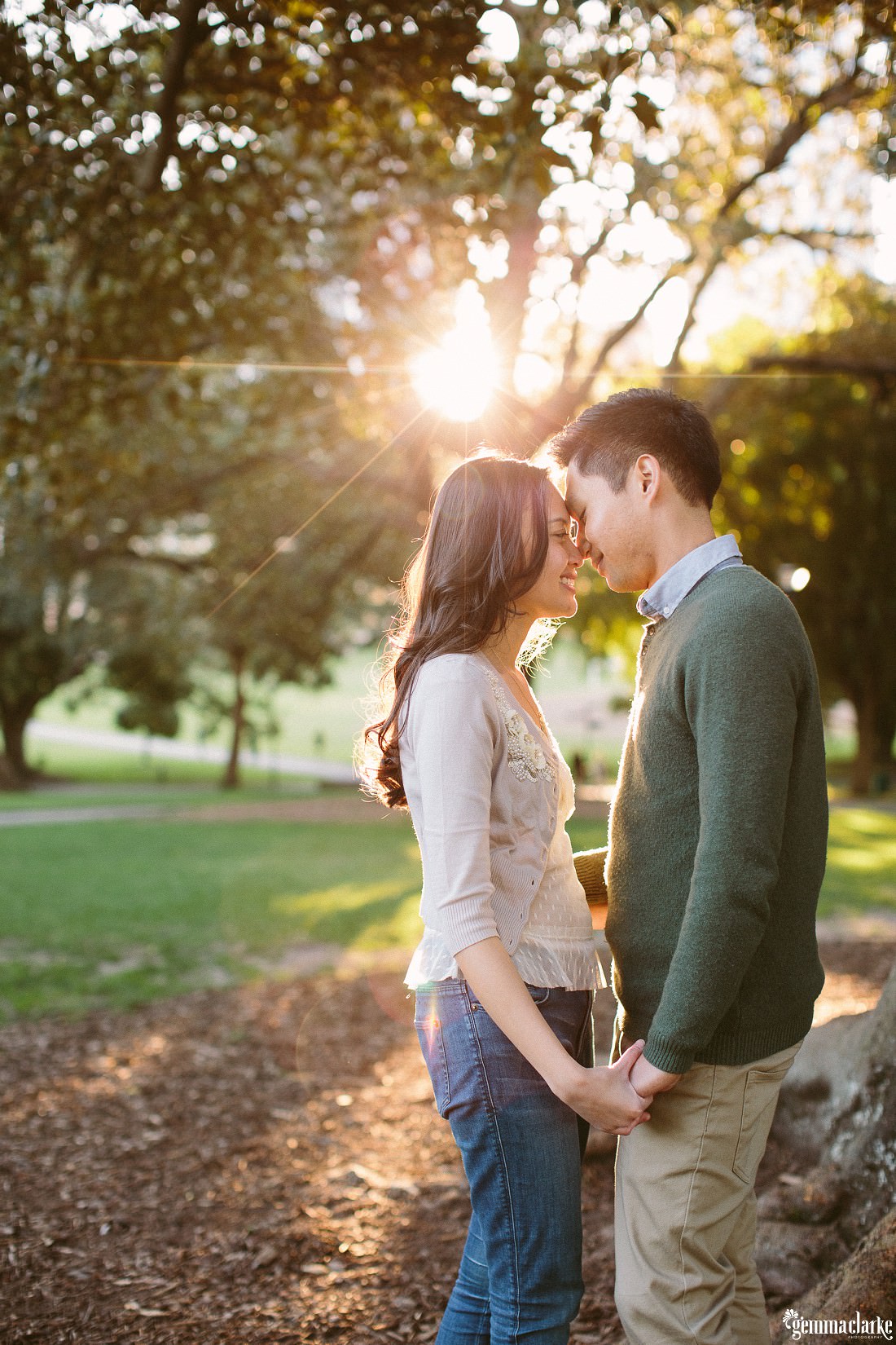 A man and woman holding hands and sharing an eskimo kiss as sunlight streams in through trees in the background - Botanic Gardens Engagement Photos