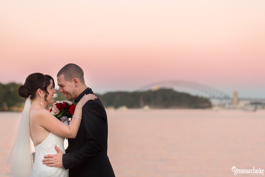 A beautiful pink sunset over the harbour bridge in the distance as the bride and groom embrace - Deckhouse Wedding