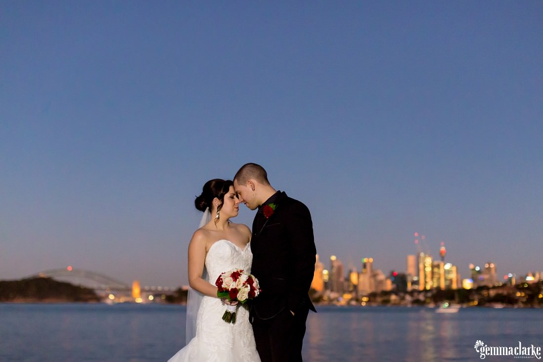 A bride and groom sharing an intimate moment with foreheads pressed together by the water with the Sydney skyline in the background - Deckhouse Wedding