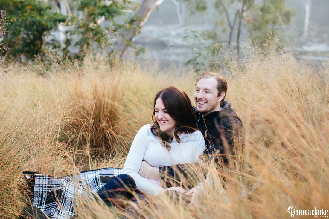 The couple are sitting on black and white tartan picnic blanket in the long yellow grass - Lake Parramatta