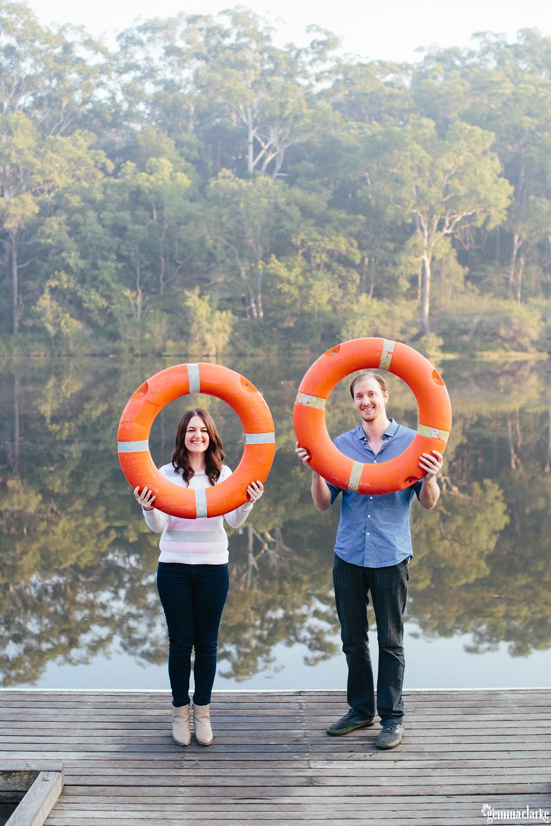 Full length photo of couple holding up bright orange lifesavers in front of their heads and smiling with the lake in the background - Lake Parramatta