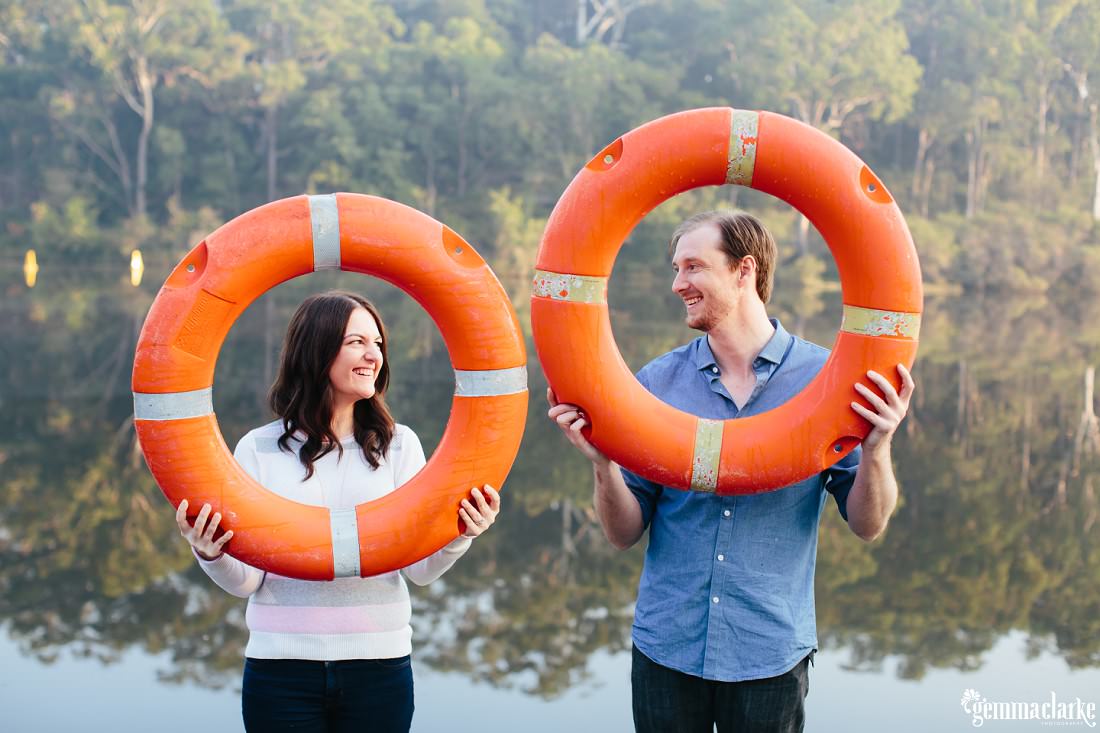 Engaged couple hold up bright orange lifesavers with their heads showing through the middle while standing in front of a lake - Lake Parramatta