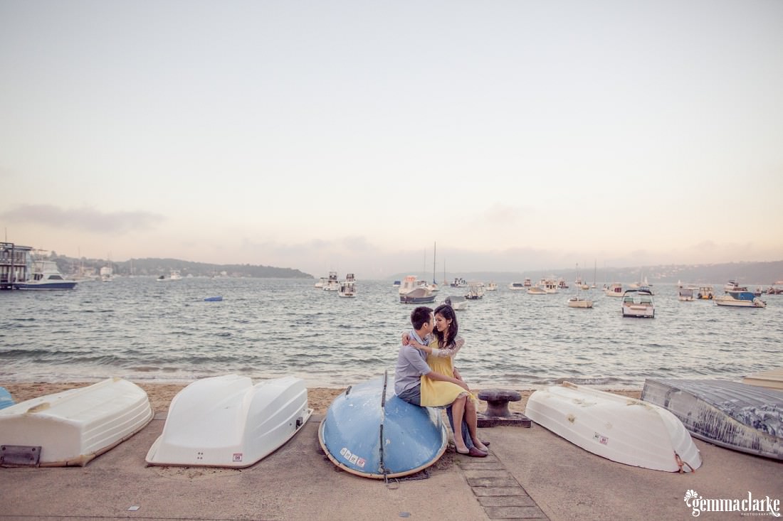 A woman sits on a man's lap as they embrace while sitting on an upturned boat on a beach - Watsons Bay Engagement Photos