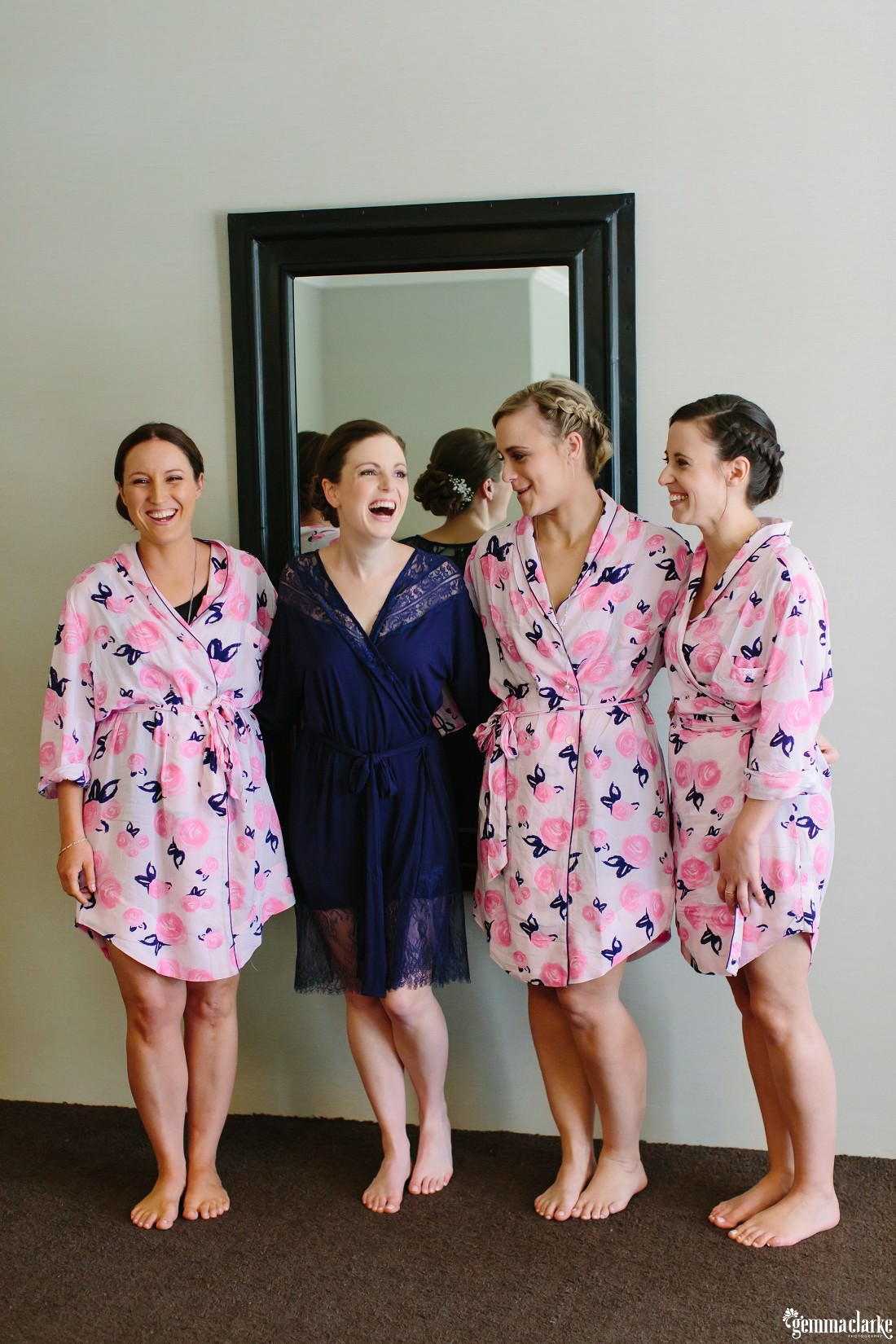 A bride and her bridesmaids in robes, smiling in front of a mirror