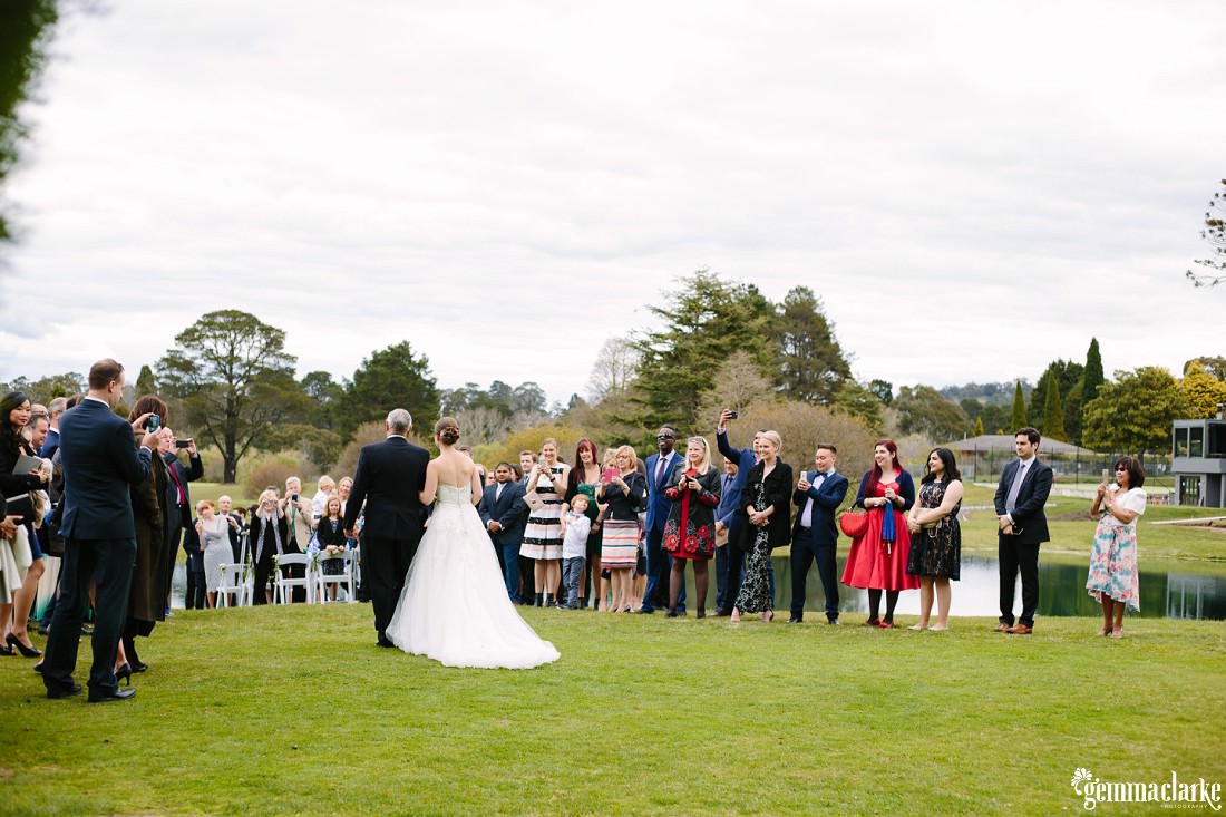 Wedding guests look on and take photos as the bride is walked down the aisle by her father