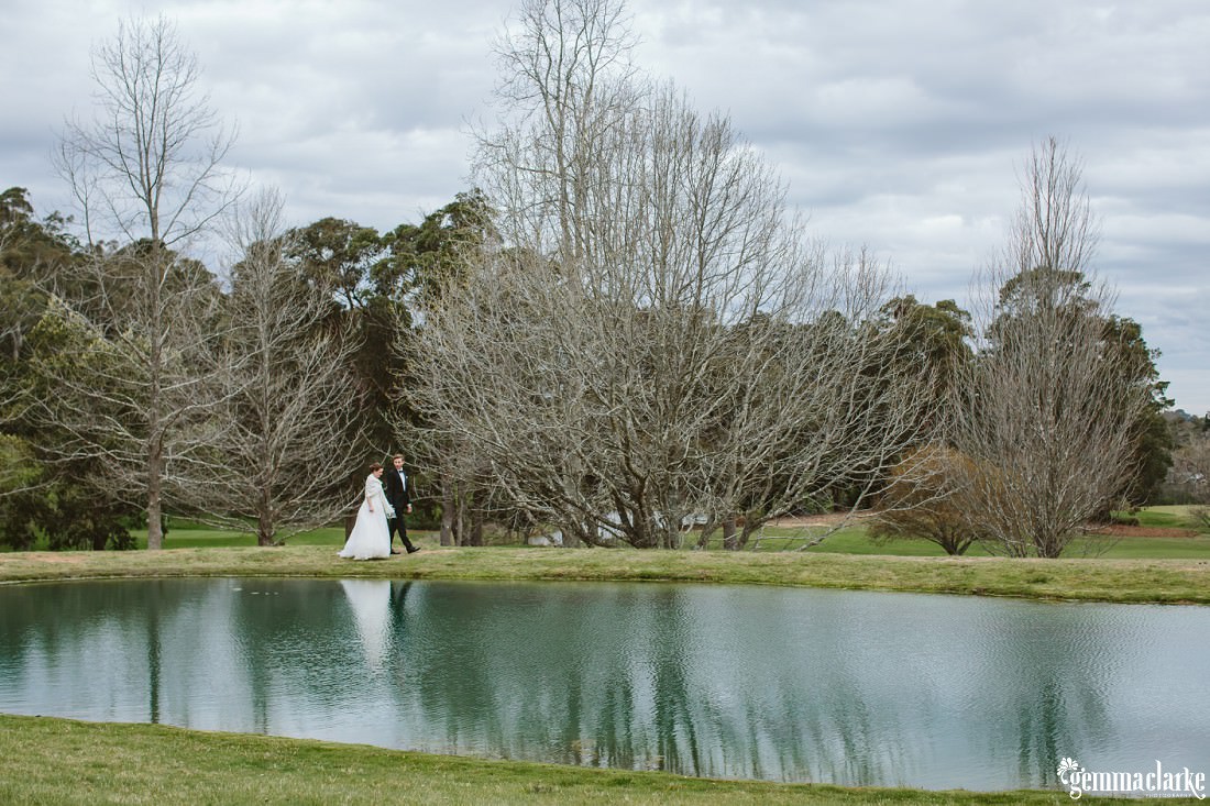 A bride and groom holding hands and walking alongside a small lake