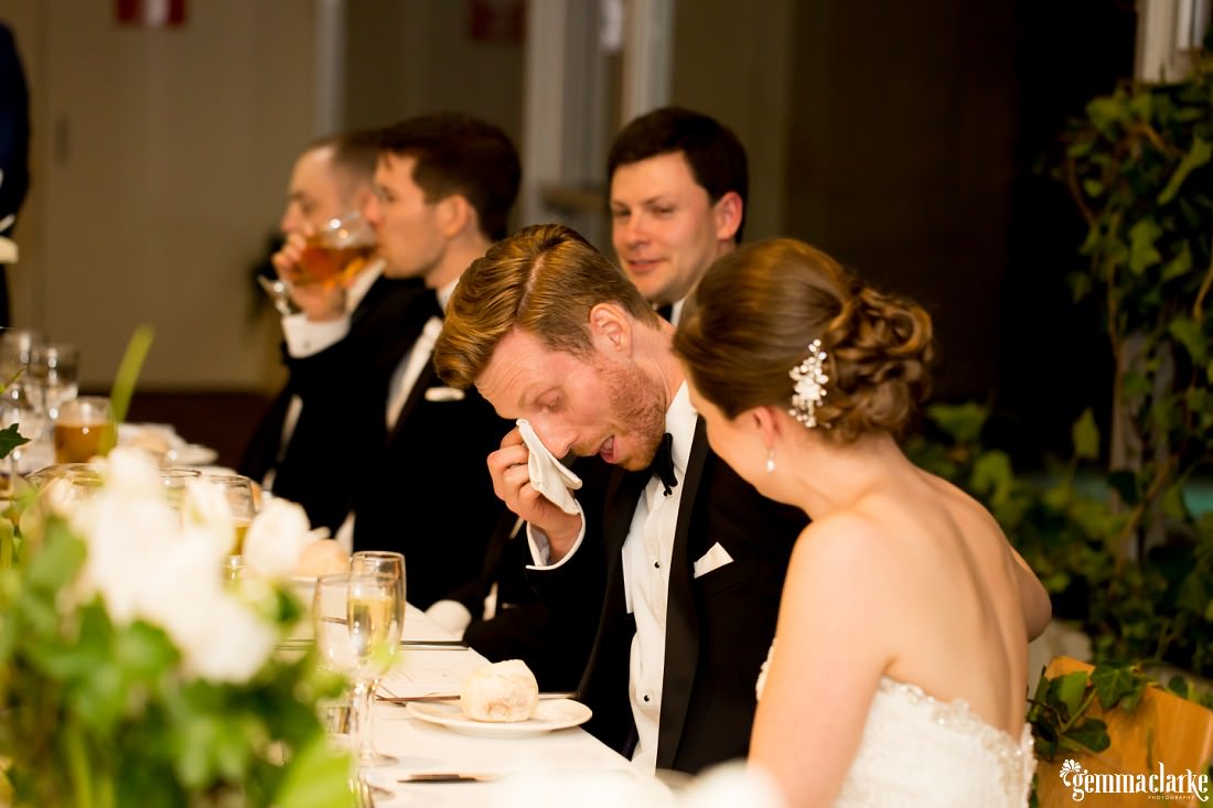 A groom wipes a tear from his eye at his wedding reception