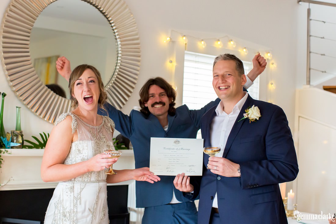 A smiling bride and groom proudly display their marriage certificate as their celebrant raises his arms in celebration