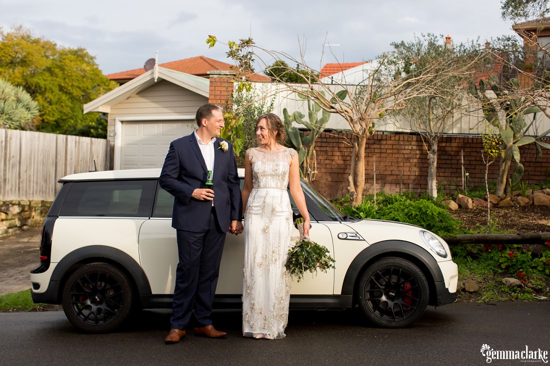 Bride and groom holding hands while standing in front of the groom's Mini countryman car, he is holding a beer and she is holding a native bridal bouquet at this Wedding at Home