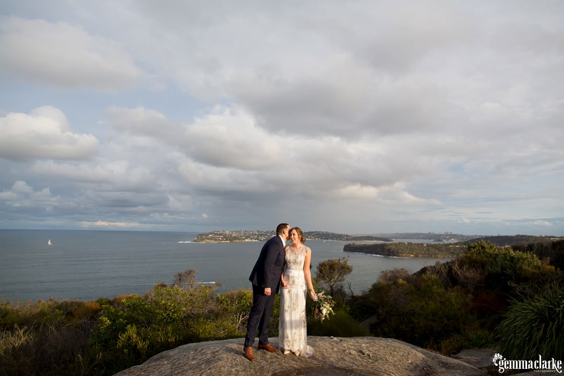 A bride and groom standing on a rock with a magnificent view of the ocean and coastline in the background