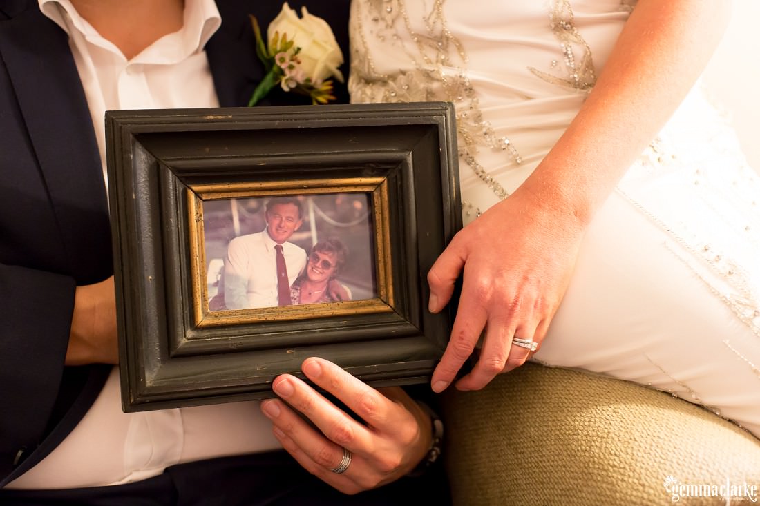 A bride and groom holding a framed photo of the groom's parents