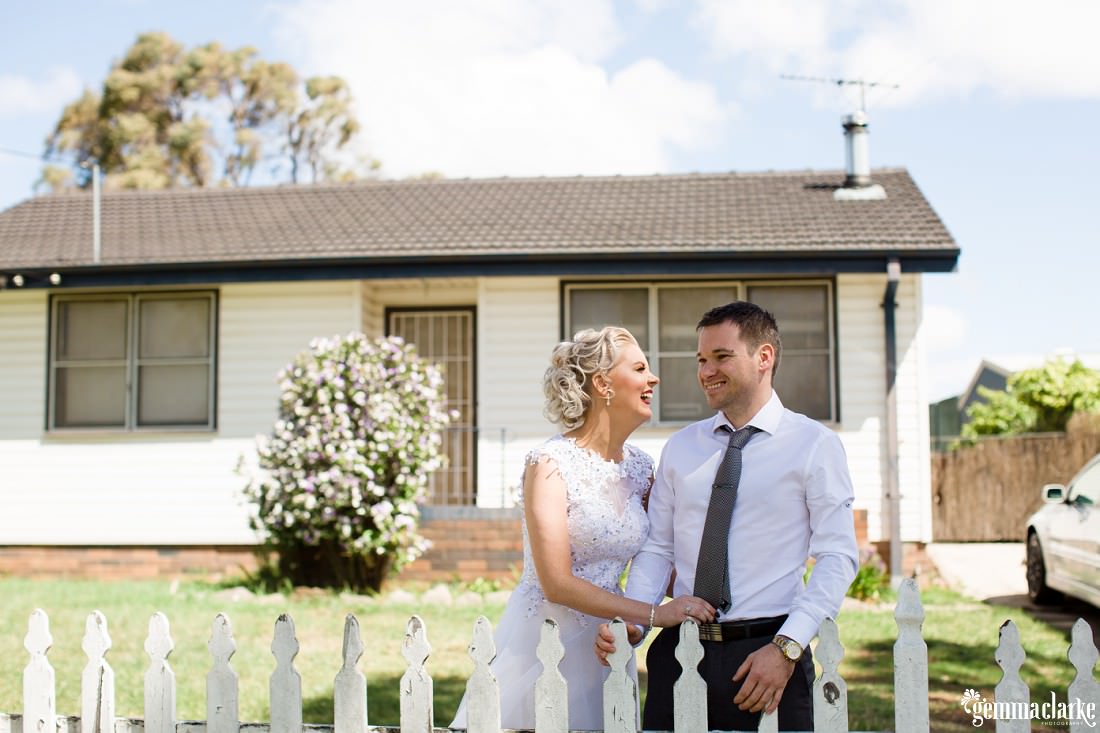 A bride and groom standing in front of their house with white picket fence
