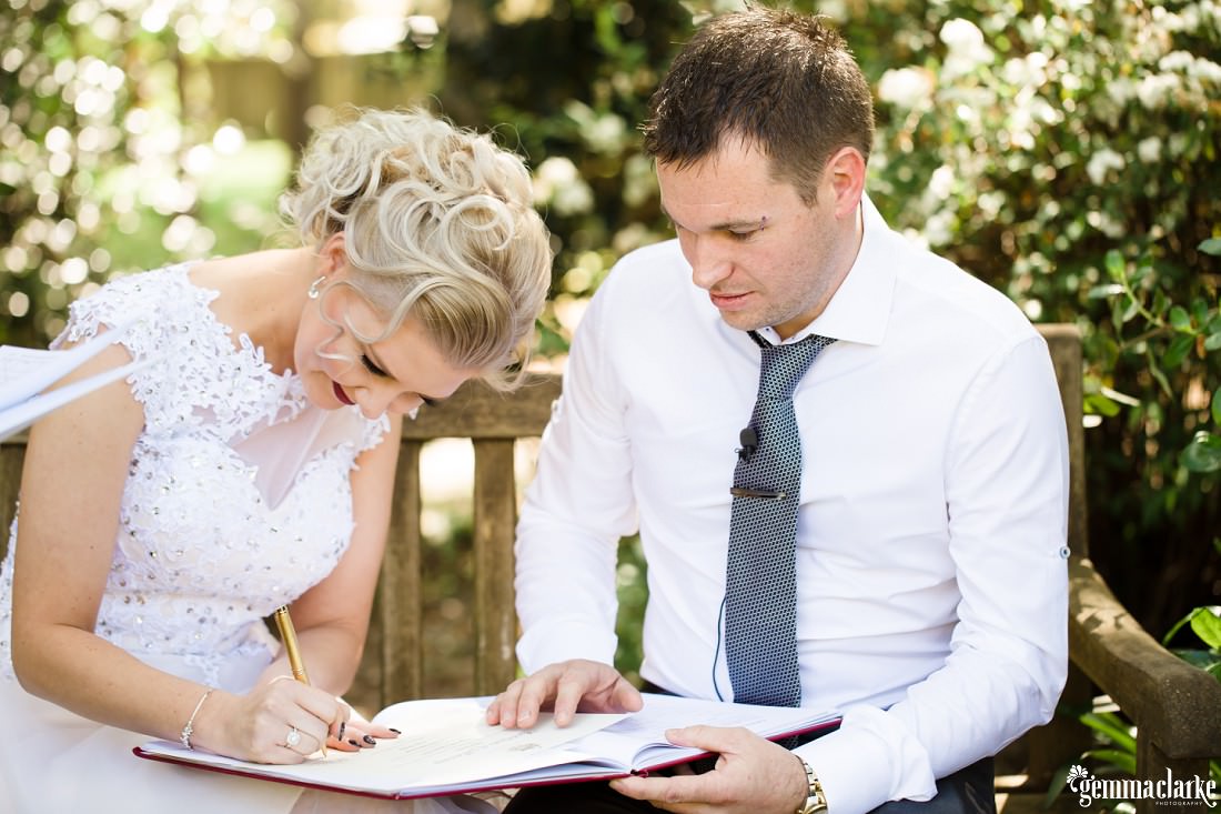 A bride and groom sitting on a wooden bench signing their wedding papers