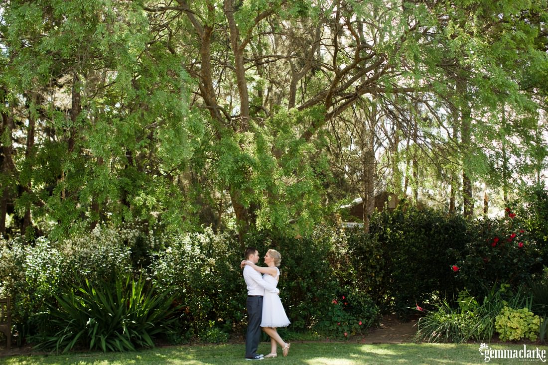 A bride and groom with their arms around each other under a large tree in a garden