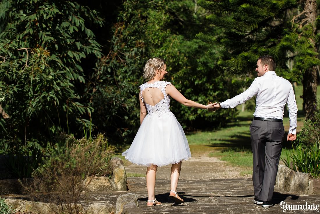 A bride and groom looking at each other and holding hands while walking down a path through a garden