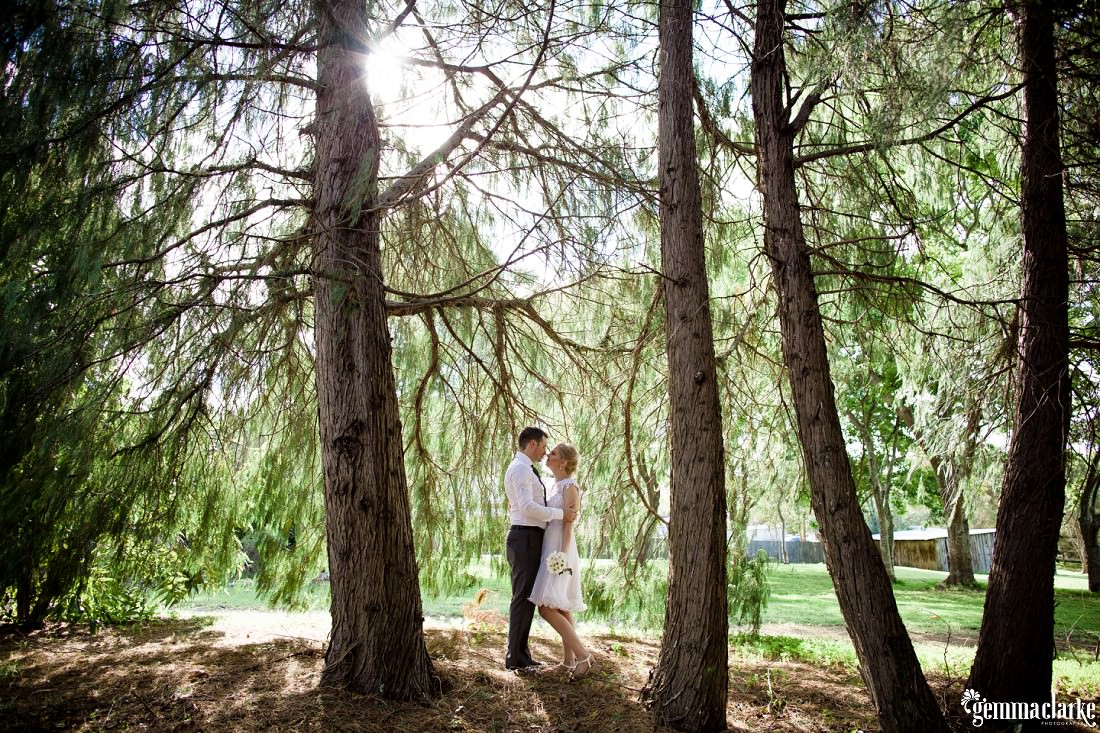 A bride and groom stand close among trees with sunlight streaming in from behind