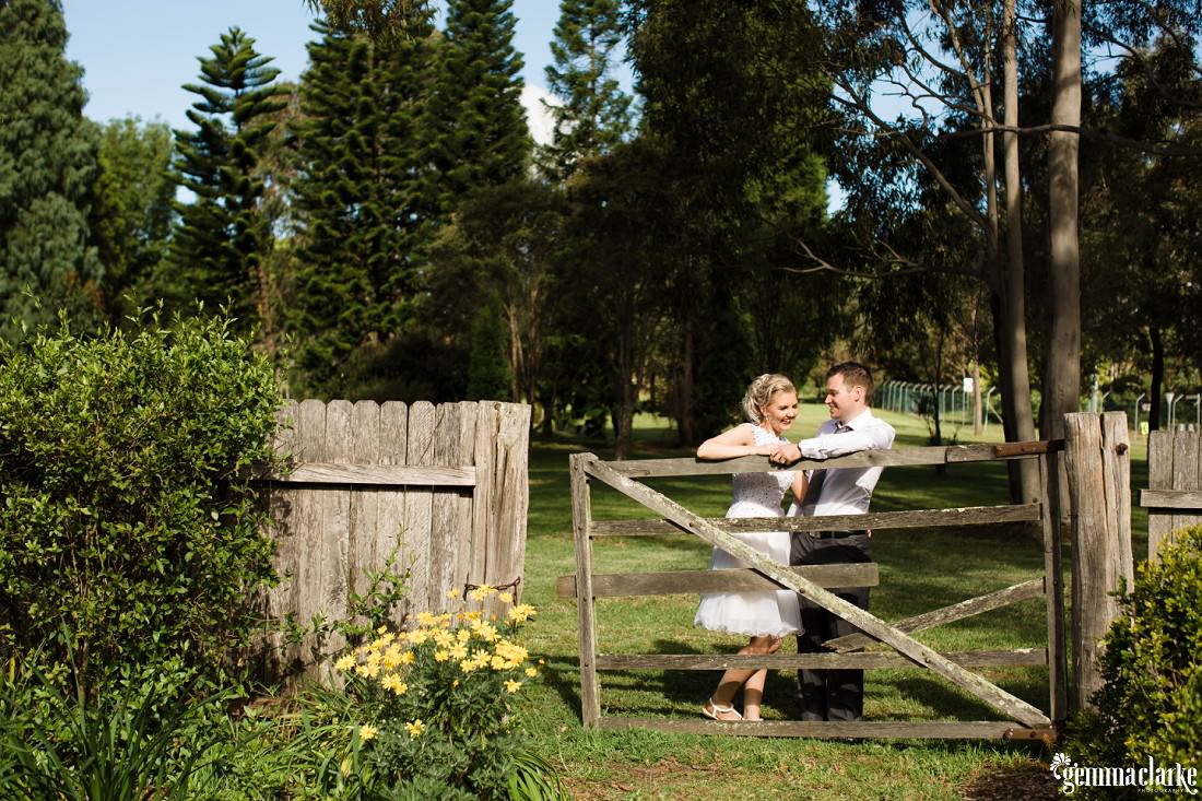 A smiling bride and groom together leaning on an old wooden gate in a park