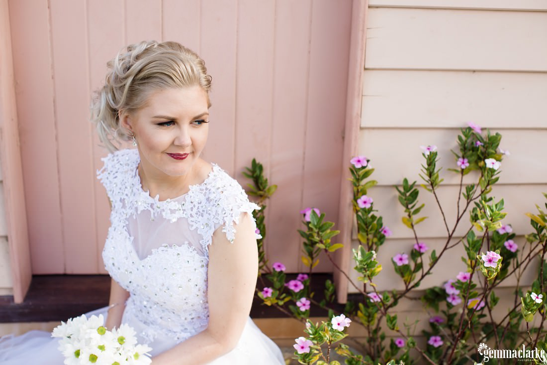 A smiling bride sitting on a step in front of a pale pink door and some white and purple flowers