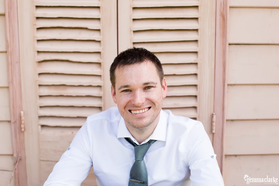 A smiling groom sitting on a step in front of a pale pink door