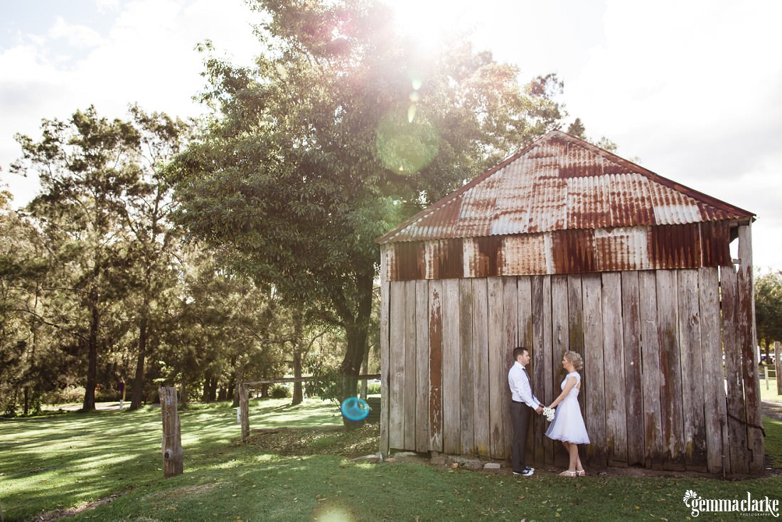 A bride and groom standing together and holding hands in front of an old wood and corrugated iron structure