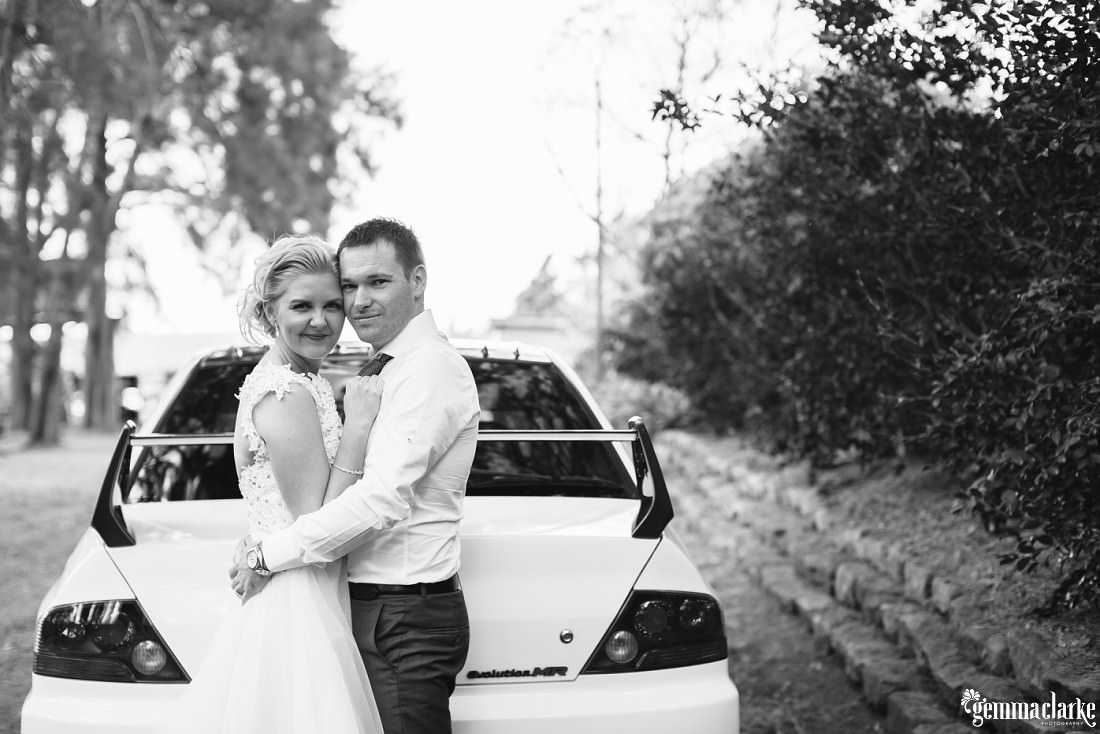 A bride and groom standing close in front of the back of a car