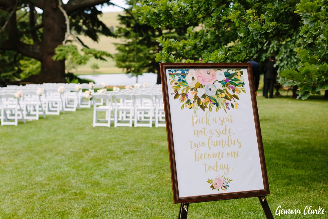 Set up of a ceremony on green grass with the big trees in the background, There is a sign in the foreground that tells the guests to pick a seat, not a side as we become one today at this Bendooley Estate Wedding