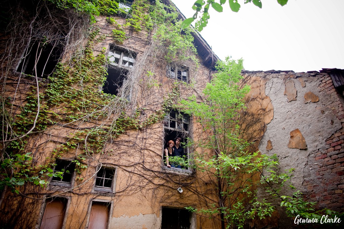 An amazing abandoned yellow building with peeling paint and vines growing over it with the couple peeking out from one of the windows in these Berlin couple portraits
