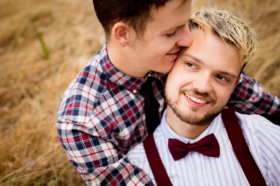Male couple snuggle on the grass while sitting up.