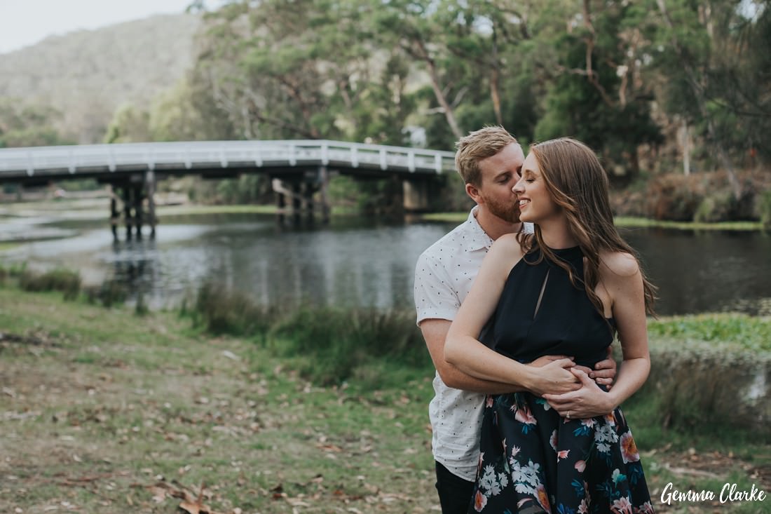 A couple cuddle by the riverbank with the white bridge in the background at Aduley at these sydney engagement photos