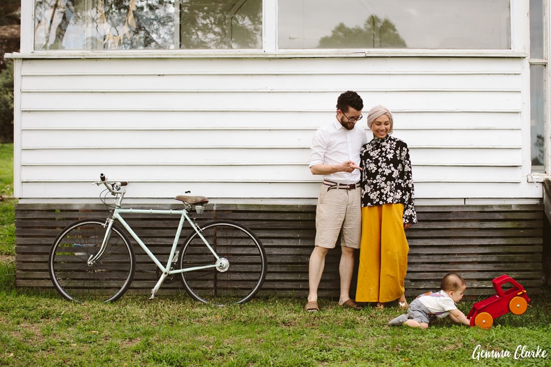 Couple stand against a weatherboard house with bike and baby pushing a vintage red car at these Family Lifestyle Portraits 