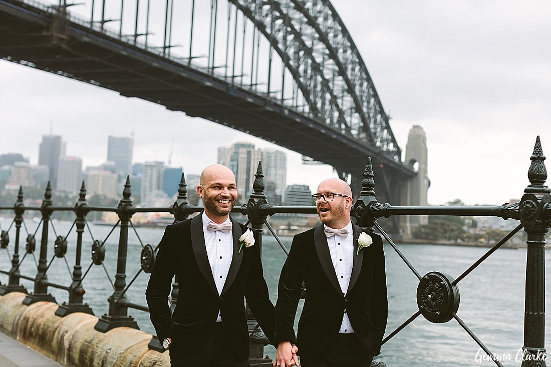 Two grooms walk alongside the harbour hand in hand with the Harbour Bridge in the background at this Sydney Gay Wedding