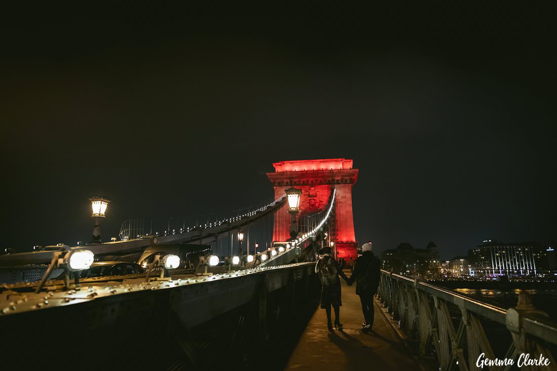 The famous bridge in Budapest at night lit up red as the couple walk hand in hand along the walkway