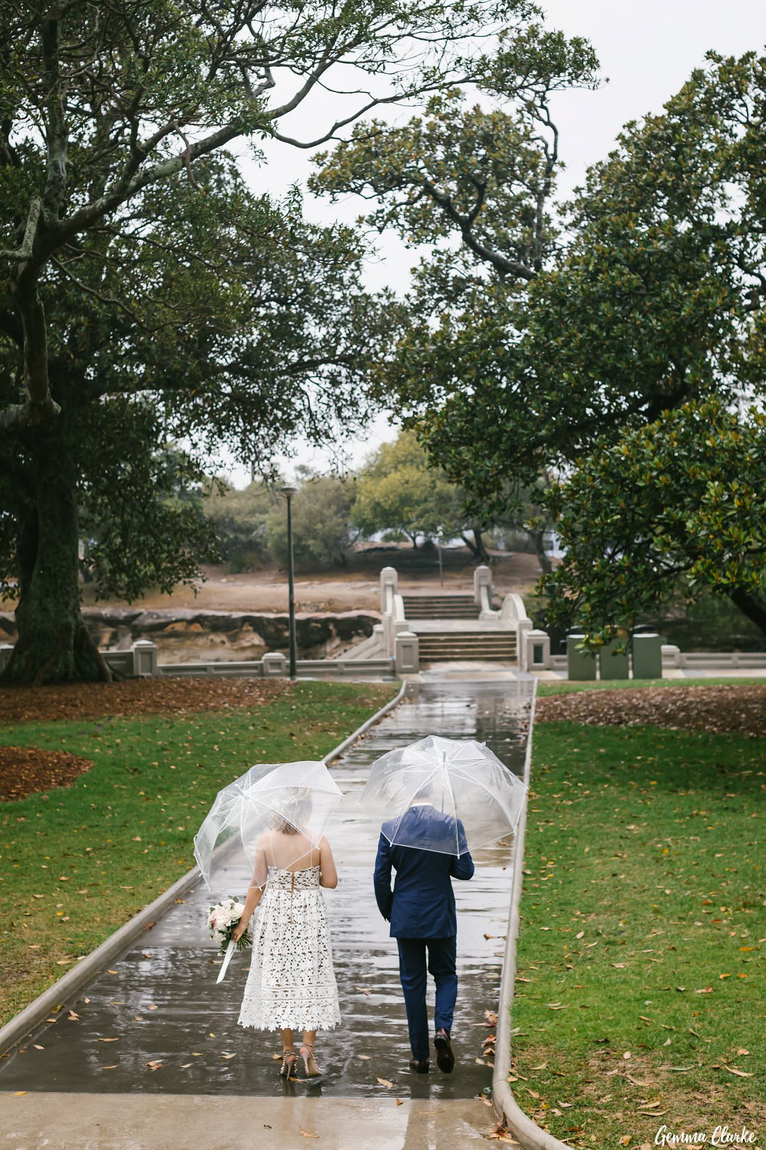 Bride and groom walk with clear umbrellas down the path at Balmoral Beach after their ceremony - rain on your wedding day